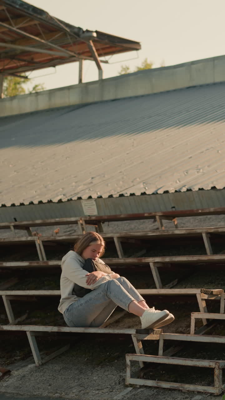 dama sentada sola en las gradas del estadio en una postura reflexiva, manos sosteniendo una bolsa, el fondo muestra el techo de metal del estadio, árboles y vegetación
