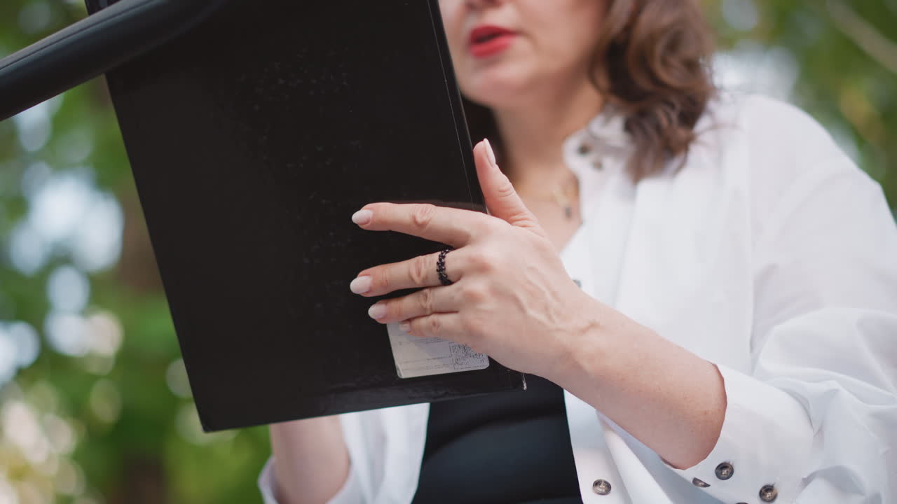 Caucasian woman holding decree document closeup of hands and rings, black folio on clipboard, presenting mandate to audience, formal tone, outdoor garden backdrop, authoritative lifestyle moment
