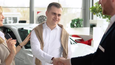 male dealer passing car keys to happy couple and shaking hand of satisfied client. auto business, car sale, technology and people concept
