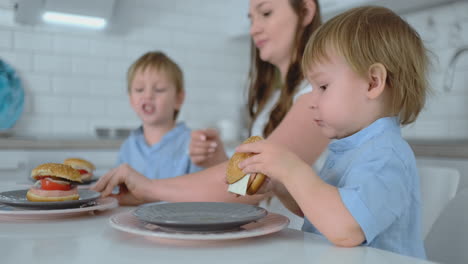 baby boy sitting in the kitchen with his mother and brother eating a burger and smiling. healthy food, home burgers