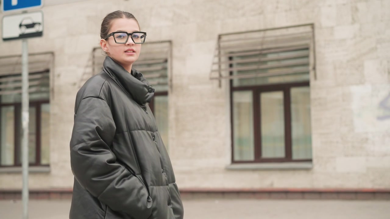 Close up of woman with glasses and tied hair standing outdoors in front of urban building facing away from camera. Woman wears black padded jacket as woman remains stationary throughout