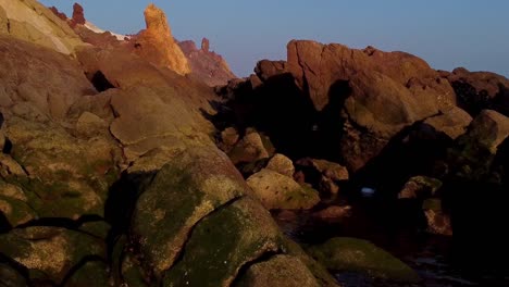 Fly-over-dark-black-sea-coastal-beach-rocky-mountain-polished-by-wind-erosion-in-background-and-sunset-golden-light-on-scene-the-blue-wave-and-surfing-club-concept-campsite-marine-lodge-resort-in-Iran