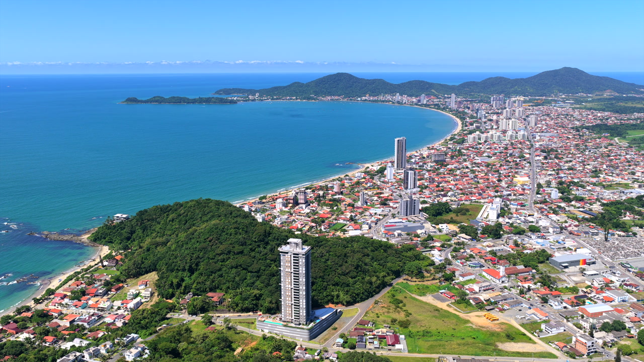 Drone view of Penha, Santa Catarina, Brazil, with Praia da Armação do Itapocorói, Ponta da Vigia headland, coastal skyline and Serra do Mar mountains in background.