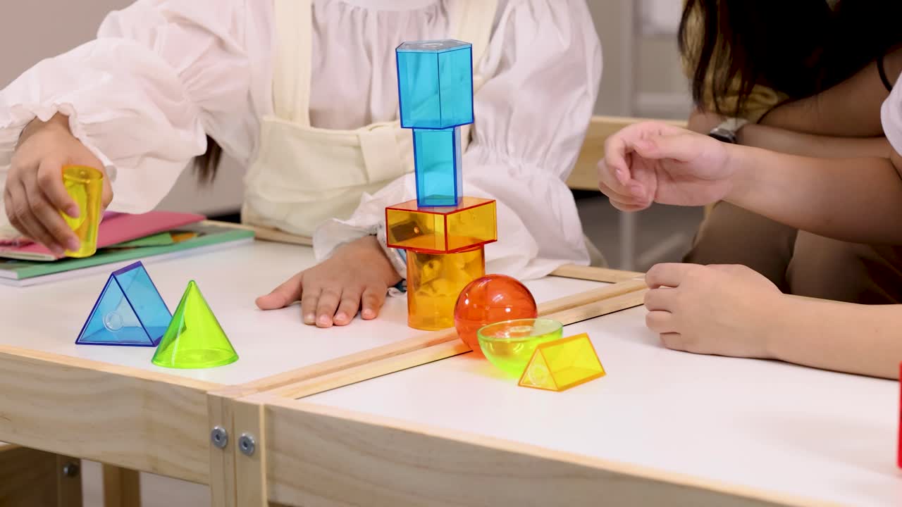Kids collaboratively stack translucent geometric blocks on classroom table, learning balance, shapes, and colors