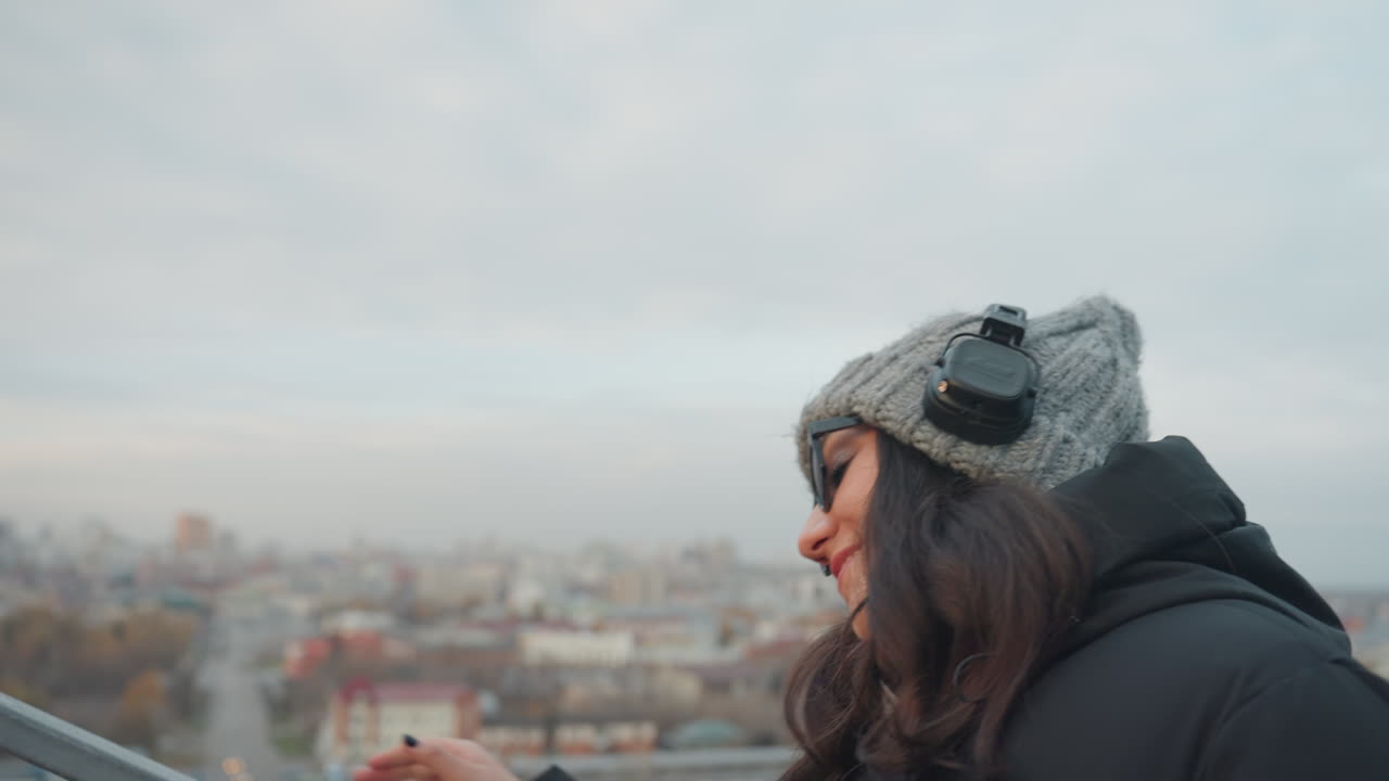 Donna con cappello di lana e cuffie si gode la vista dello skyline annuendo a ritmo di musica, il primo piano del profilo mostra un sorriso e un movimento rilassato, un sottile ritmo di danza e un'espressione calda sullo sfondo di un panorama urbano.