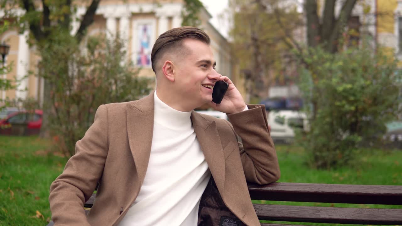 Elegant smiling man talking on the phone while sitting on a bench in the street
