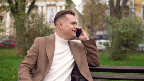 elegant smiling man talking on the phone while sitting on a bench in the street