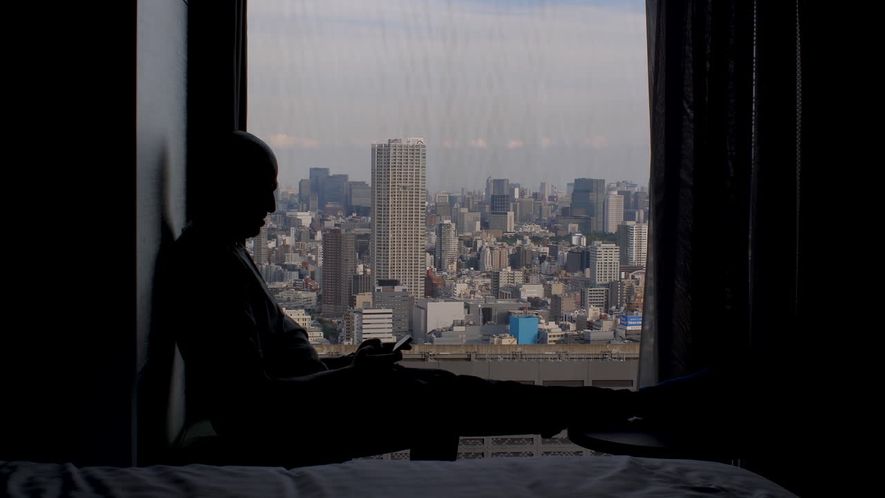광활 한 도시 대도시 전망 을 가진 휴대 전화 를 사용 하여 창문  에 앉아 있는 남성 의 실루