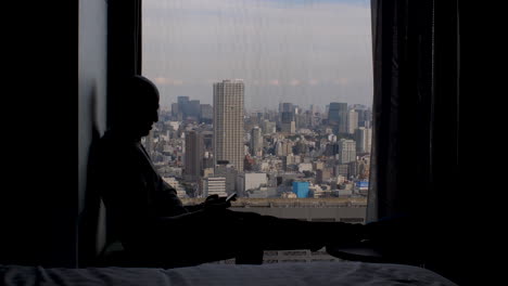 Silhouette-Eines-Mannes,-Der-Neben-Dem-Fenster-Sitzt-Und-Ein-Mobiltelefon-Benutzt,-Mit-Weitem-Blick-Auf-Die-Metropole