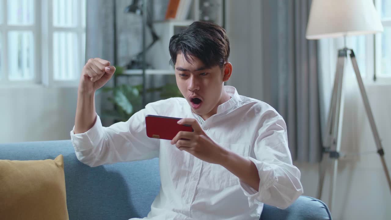Asian Man Celebrating Winning Game On Smartphone While Lying On Sofa In The Living Room