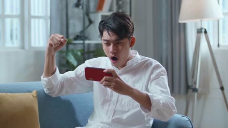 asian man celebrating winning game on smartphone while lying on sofa in the living room