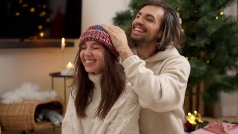 Happy-brunette-guy-puts-a-red-woolen-hat-on-his-brunette-girlfriend-in-a-white-sweater-hugs-her-and-rejoices-in-a-cozy-room-decorated-in-Christmas-style-with-a-Christmas-tree