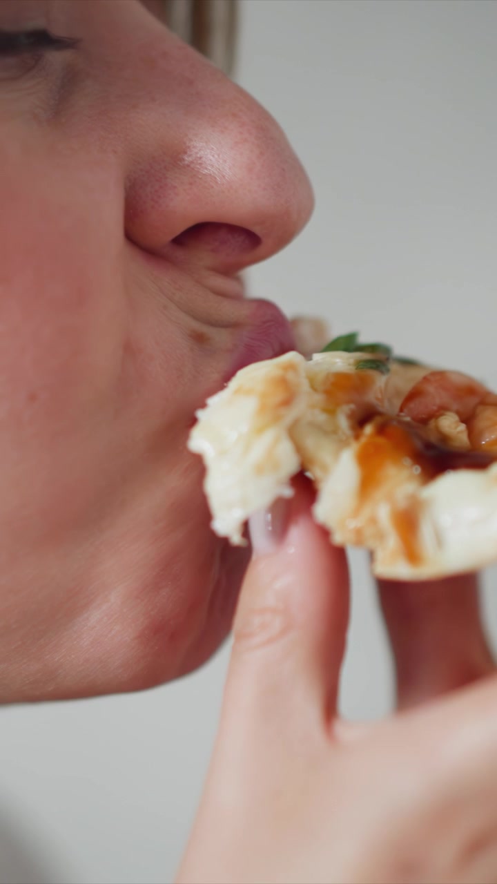 Close up of woman eating pizza. subject holds and takes bites from food with fine details like sauce texture on crust. Background is plain white to emphasize action