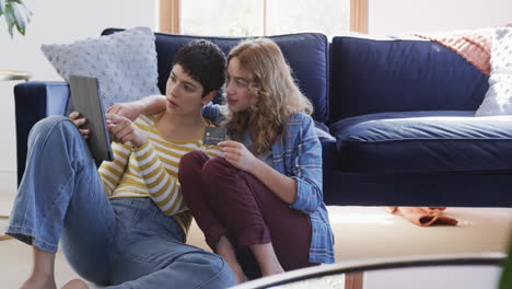 happy caucasian lesbian couple embracing on floor, using tablet and credit card in sunny house