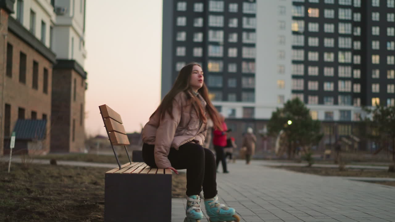 A girl in a peach jacket and black trousers is seen rollerblading towards a park bench in an urban environment. As she sit and rest, other people can be seen in the background