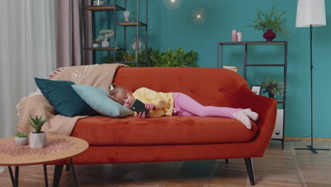 Child toddler hold smartphone watching online cartoons, chatting with friends on sofa alone at home
