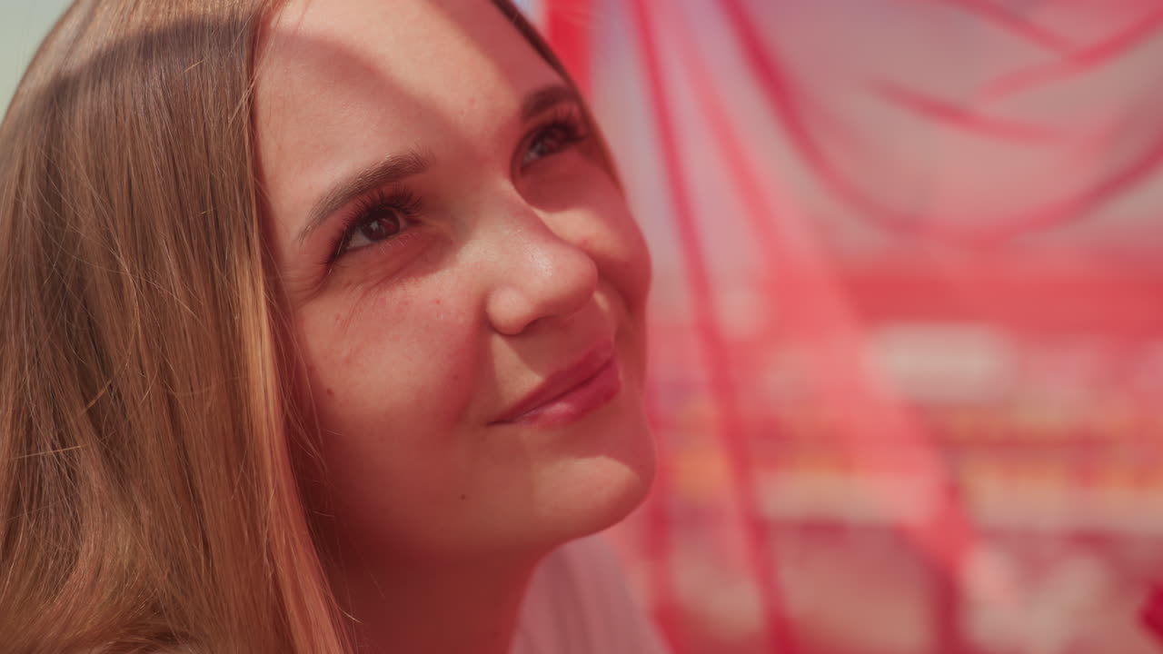 Eine lächelnde Frau vor roten Vorhängen, die das Sonnenlicht auf ihrem Gesicht einfängt, eine sanfte Nahaufnahme, die einen hoffnungsvollen Blick und ein schüchternes Lächeln bei einem romantischen Date auf dem Dach einfängt, warme Töne, geringe Schärfentiefe, natürliches Make-up und Gelassenheit