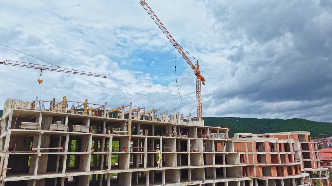 Construction site with cranes and unfinished building under cloudy sky in mountain area at midday