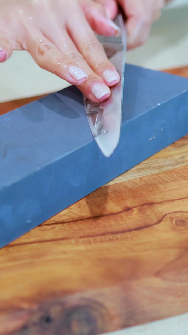 Hands methodically sharpen a stainless steel chef’s knife on a rectangular whetstone, using smooth, controlled strokes in bright, natural lighting