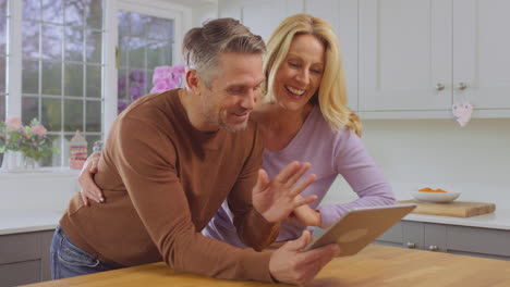 mature couple at home in kitchen using digital tablet to make video call to friends or family - shot in slow motion