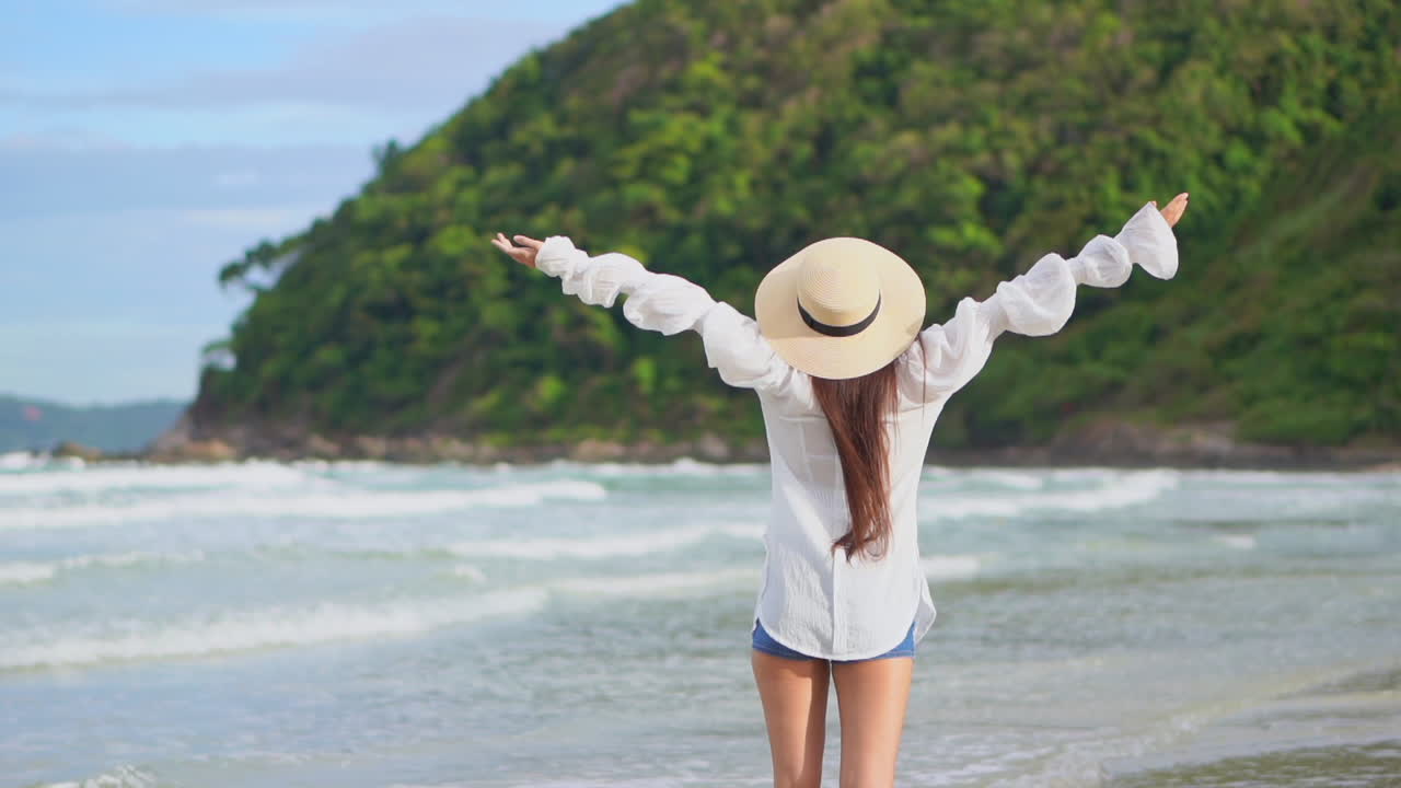 vista trasera de una chica asiática con una blusa blanca y pantalones cortos extendiéndose y levantando los brazos durante un paseo matutino en la playa de la isla, mareas marinas y montañas verdes en el fondo, a cámara lenta