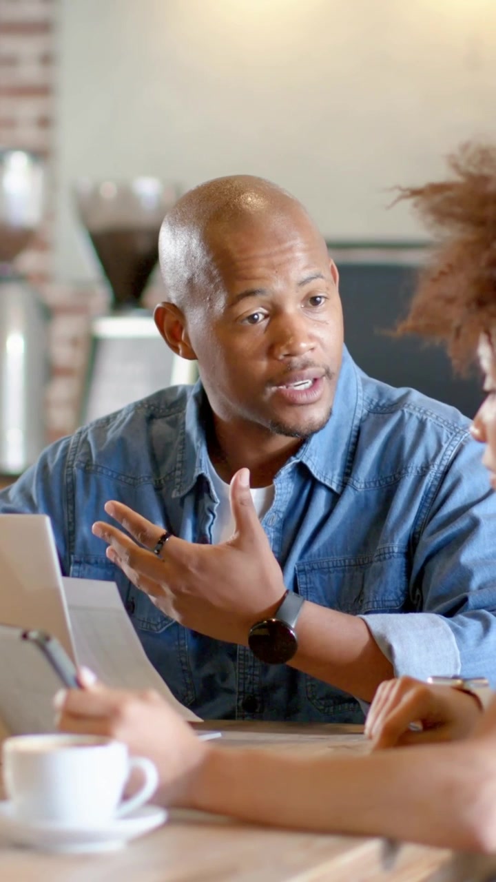 Vertical video: Talking man in denim typing on laptop at cafe planning after peer raising tablet