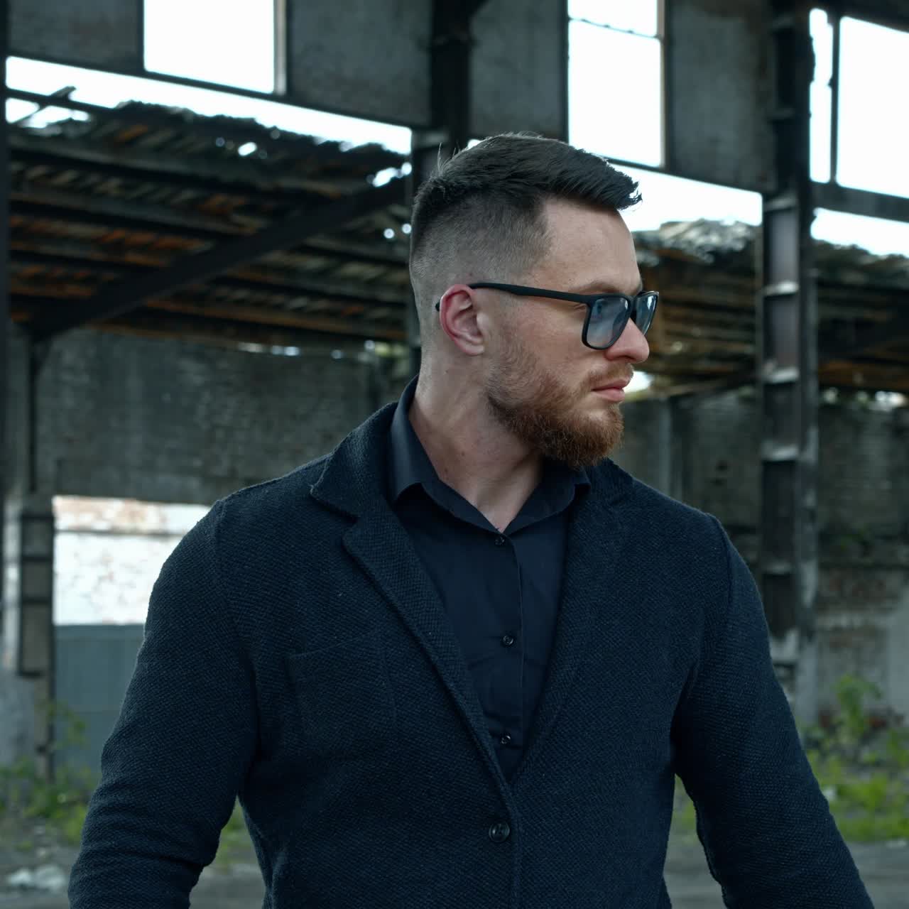 Elegant man with a baseball bat outdoors. Portrait of a serious man in stylish glasses on the background of abandoned building.