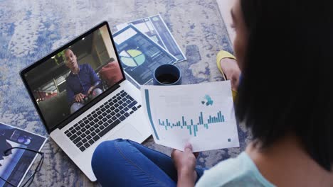 Mixed-race-businesswoman-sitting-on-floor-using-laptop-having-video-call-with-male-colleague