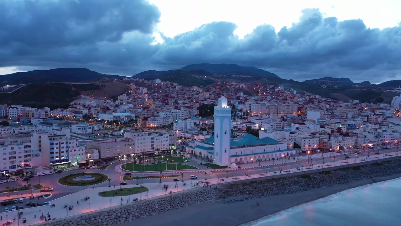 Ampia vista aerea di una città costiera mediterranea nel Marocco settentrionale all'ora blu, che mostra un lungomare illuminato, una spiaggia curva, un minareto di moschea, quartieri densi sulle colline e montagne