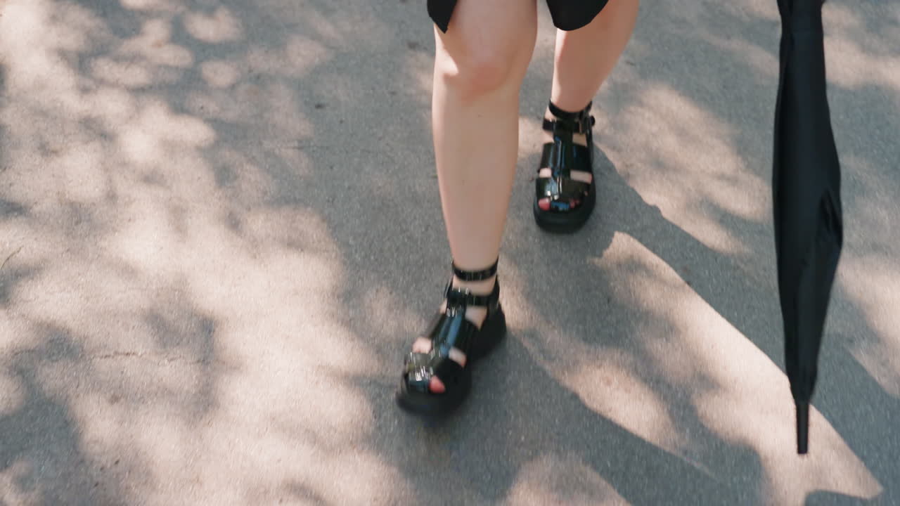 나뭇잎이 우거진 가지 아래에서 여유롭게 산책하기, 그림자가 드리워진 숲속 지역을 발로 천천히 거닐기, 그림자와 햇빛으로 둘러싸인 평화로운 공원 환경에서 우산과 샌들을 신고 편안하게 산책하기