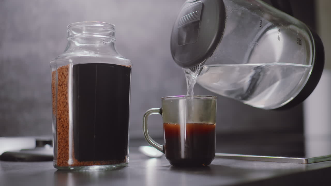 Close up of person pouring hot water from electric kettle into transparent mug containing instant coffee granules, with large coffee jar on kitchen counter