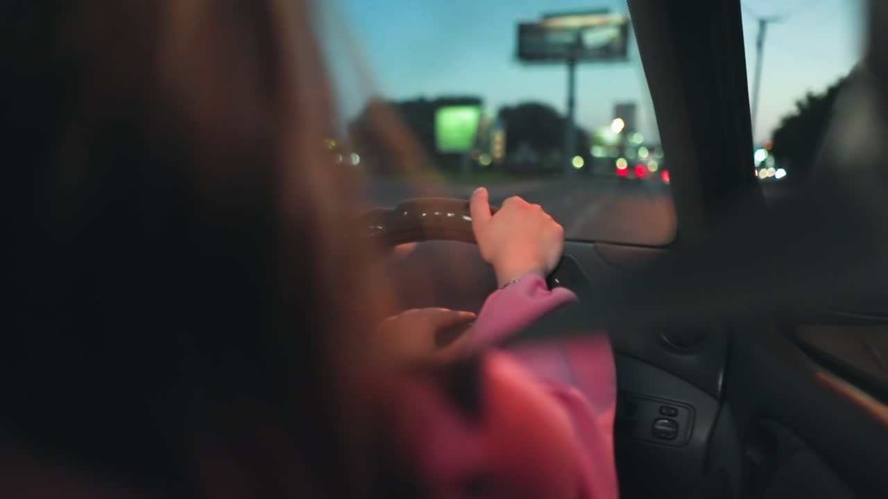 저녁에 길거리 불빛이 그녀와 도로에 반사되는 뒤에서 운전하는 여인이 보입니다. 다른 자동차는 으로 지나가는 한 차로 불빛이 약간 흐릿합니다.