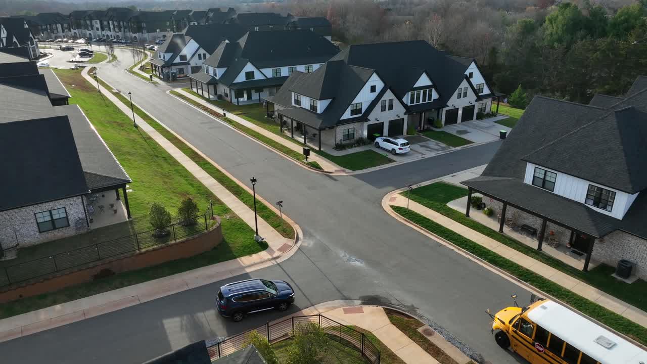 American luxury modern suburb with upscale homes, clean streets, dark gabled roofs and passing yellow school bus, surrounded by neat lawns in United States. aerial approaching shot