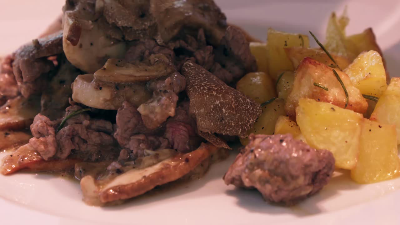 An extreme close-up shot captures someone shaving truffles onto a dish of tender meat with mushrooms and roasted potatoes, garnished with rosemary, on a white plate.
