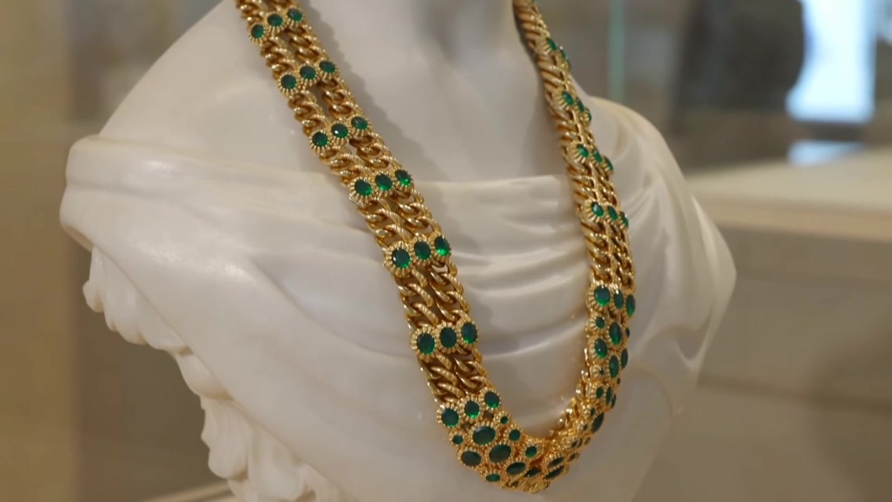 La telecamera effettua una panoramica verso il basso, mostrando un busto drappeggiato con una catena d'oro e gemme verdi in un museo.