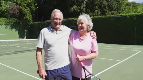 Happy-senior-caucasian-couple-holding-tennis-rackets-and-embracing-in-tennis-court