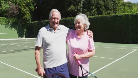 feliz pareja caucásica mayor sosteniendo raquetas de tenis y abrazándose en la cancha de tenis