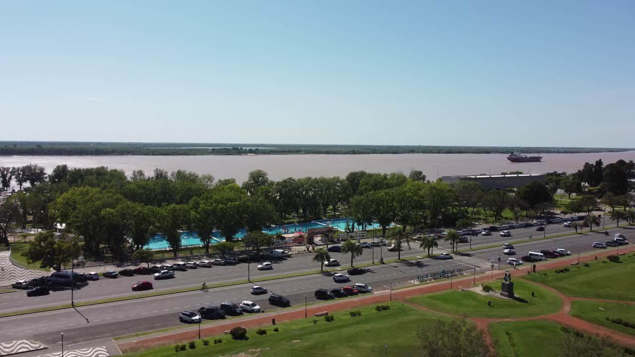 An aerial view captures a coastal avenue with car traffic and the river in the background on a sunny morning, highlighting the vibrant city life.