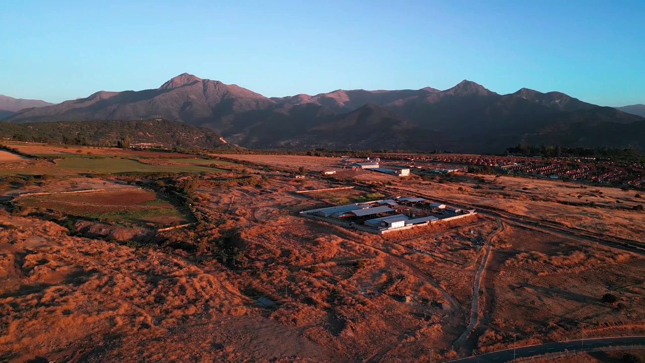 Ripresa aerea orbitale su un complesso rurale isolato in una vasta pianura arida con terra rossa e secca, strade sterrate tortuose e una spettacolare catena montuosa andina che risplende all'ora d'oro.