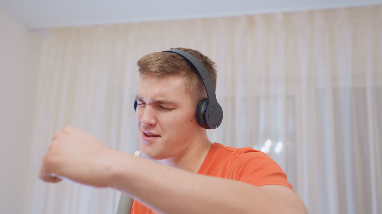 Young man in orange shirt wearing headphones sings passionately into mop handle like microphone, eyes closed enjoying rhythm, playful expression showing energy and creativity
