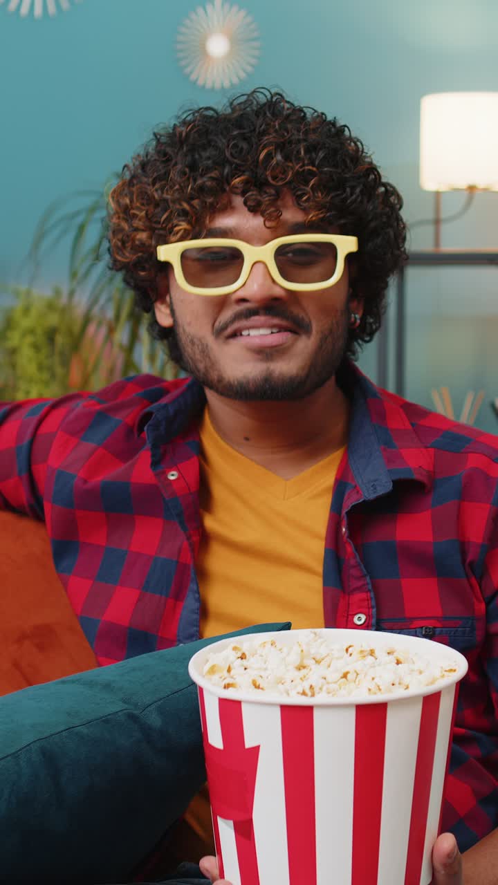 Indian man sits on couch eating popcorn and watching interesting tv serial sport game online at home