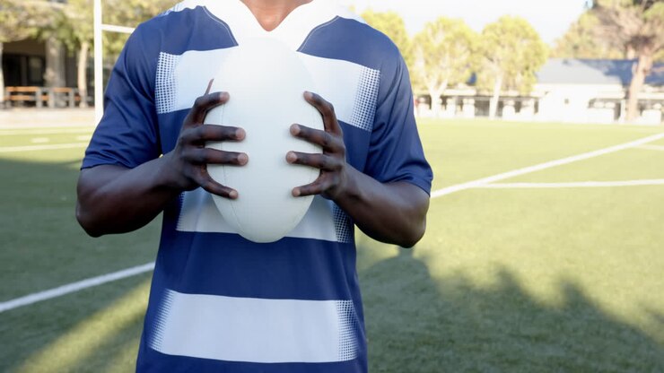 Atleta maschio afroamericano con palla da rugby in mano, con maglia blu e bianca sul campo, spazio per il testo.