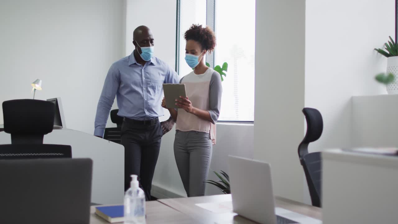 Diverse businessman and businesswoman in face masks, woman holding tablet in office