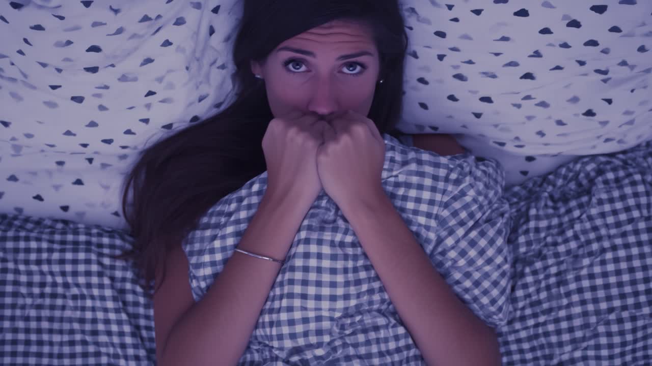 A Close-Up of a Woman in Bed, Expressing Anxiety and Fear While Gripping Her Blankets, Surrounded by Cozy but Chaotic Bedding Patterns