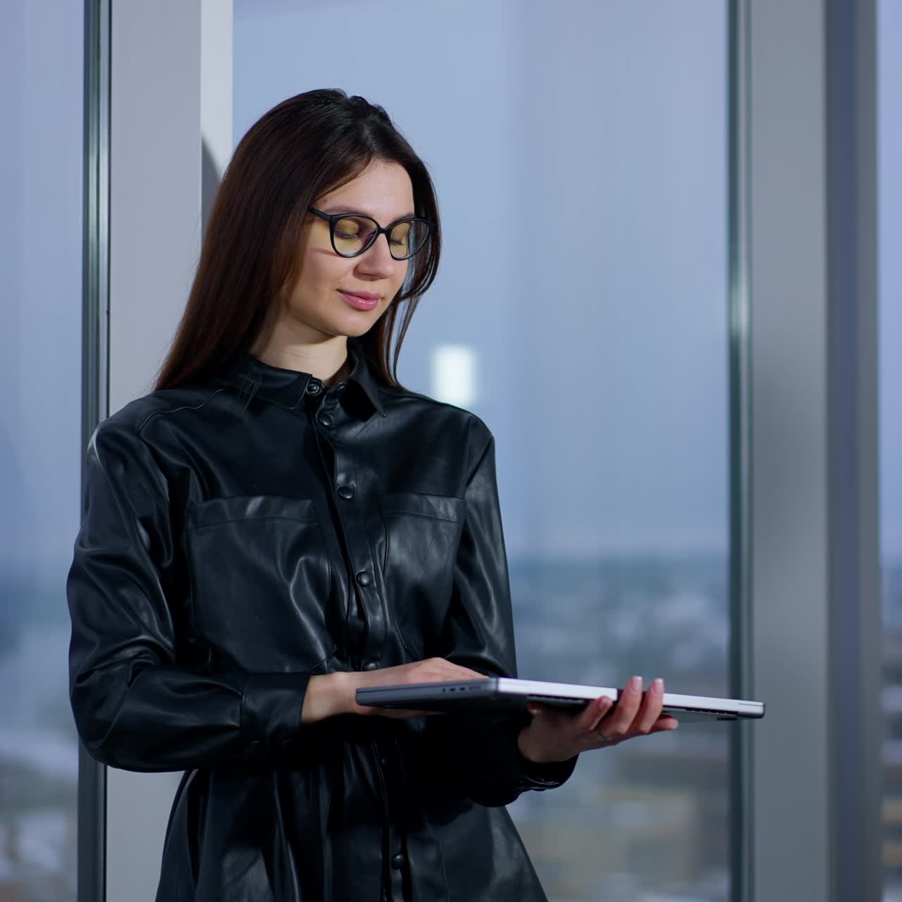 Young female business person wearing glasses types on laptop holding it in her hands. Woman finishes work, closes computer and turns to look at the window