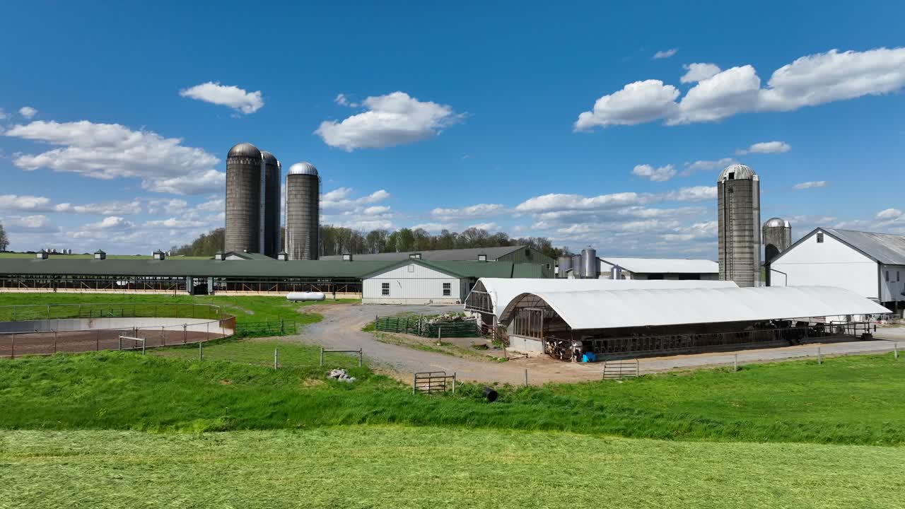 An expansive aerial view of a large dairy farm featuring multiple barns, silos, and green fields under a bright blue sky with clouds.