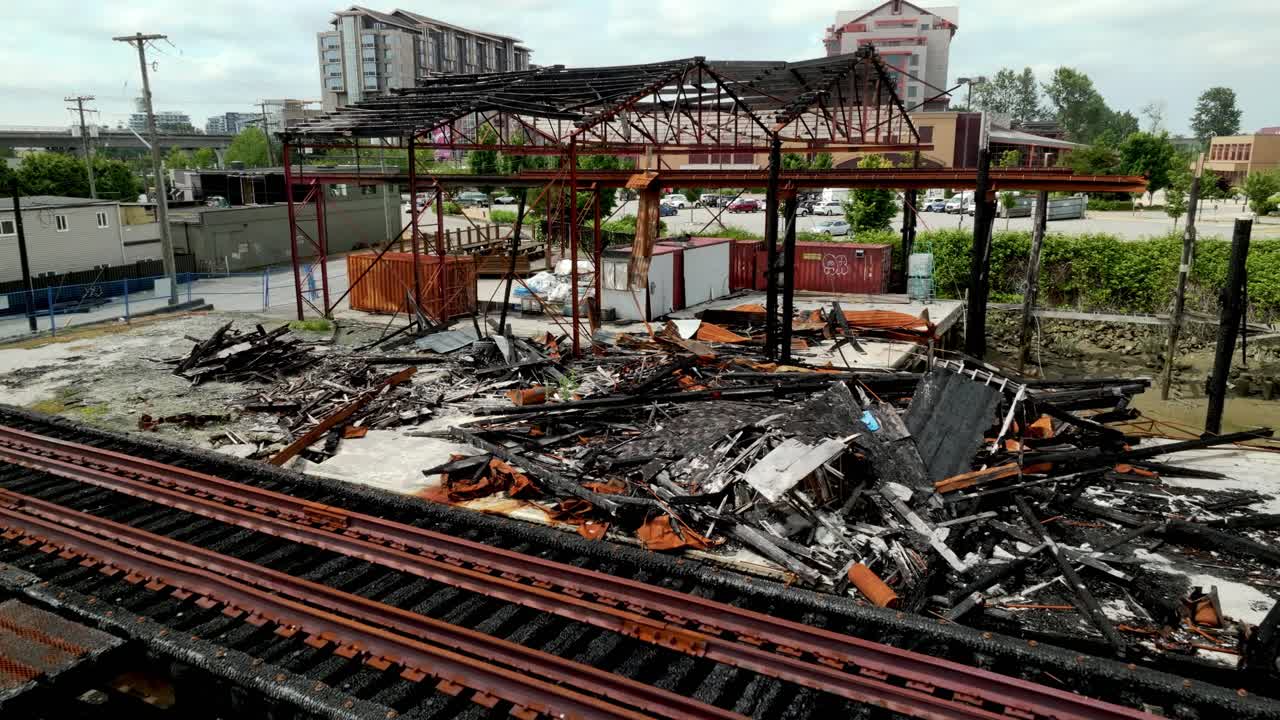 The Charred Remains of a Riverside Building Sit Beside the Rail Bridge Along River Drive in Richmond, British Columbia, Canada - Aerial Pullback Shot