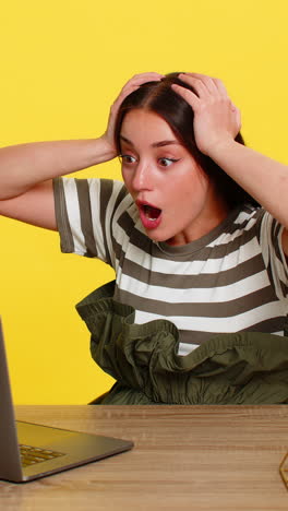 woman shocked opening mouth at table looking at laptop screen reacting to unexpected news online