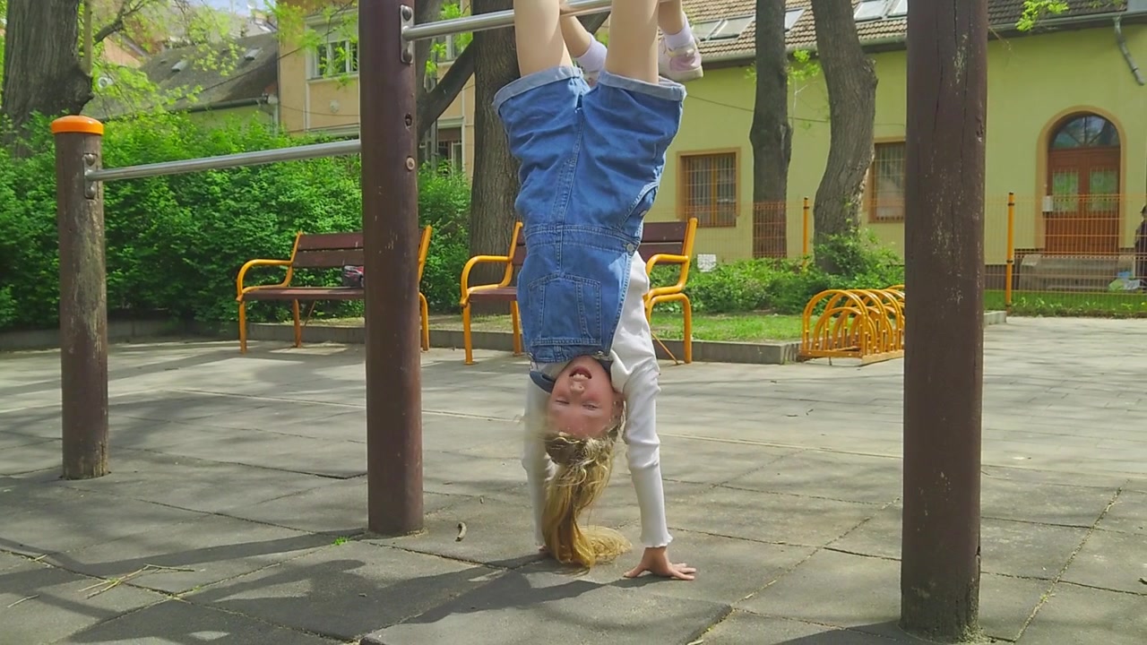 Sebuah bidikan menengah menangkap seorang gadis muda yang ceria mengenakan baju kodok denim, tersenyum sambil bergelantungan terbalik dari palang taman bermain logam saat melakukan trik handstand terbalik, menikmati permainan rekreasi di luar ruangan.