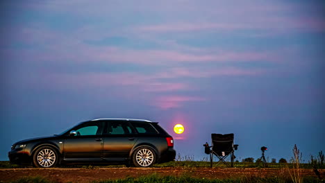 silhouette of a car and a vibrant sunset sky, with deep blue and orange hues as moon rises over hazy cloudy evening sky